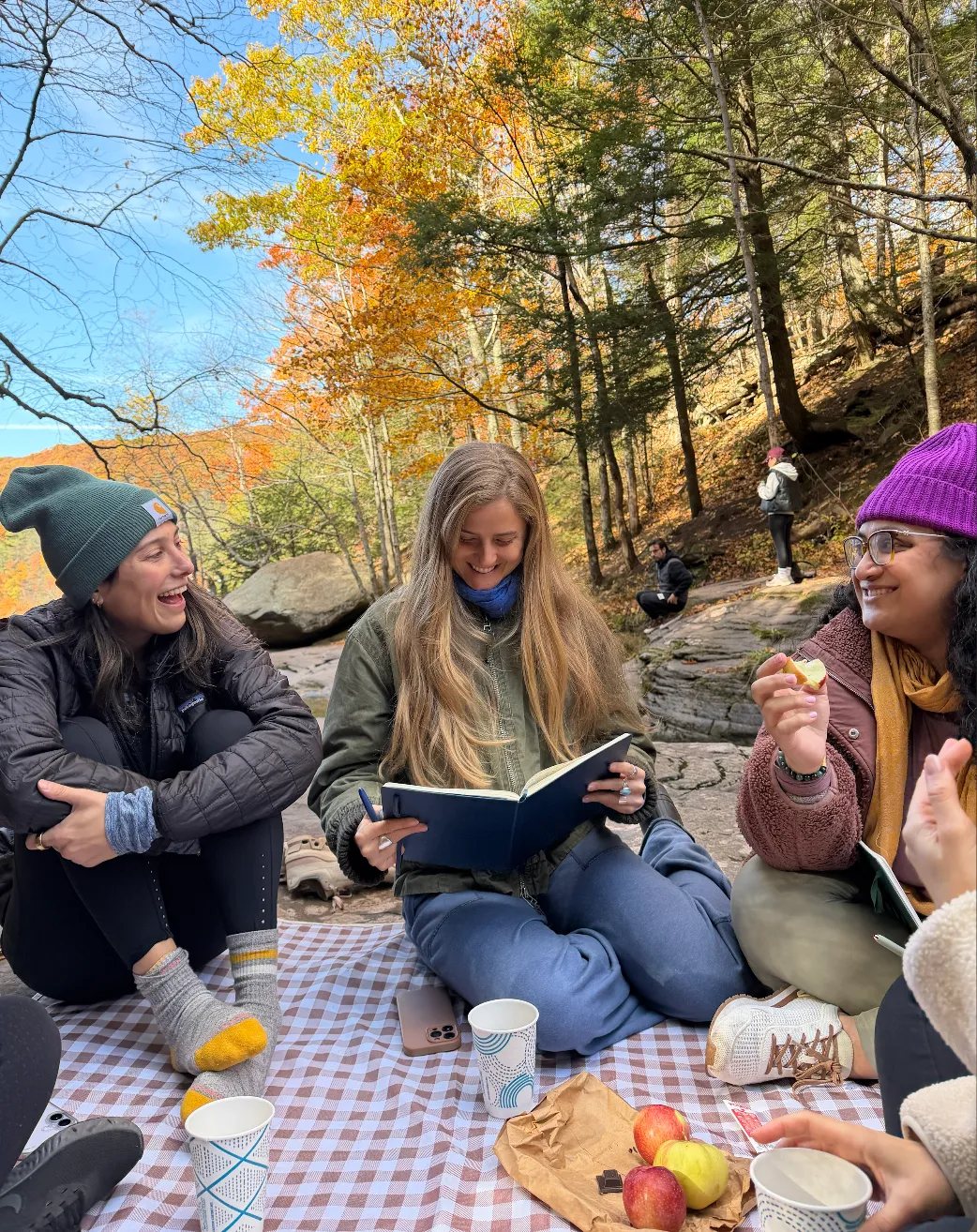 Women laughing and journaling on a picnic blanket under fall trees in the Hudson Valley
