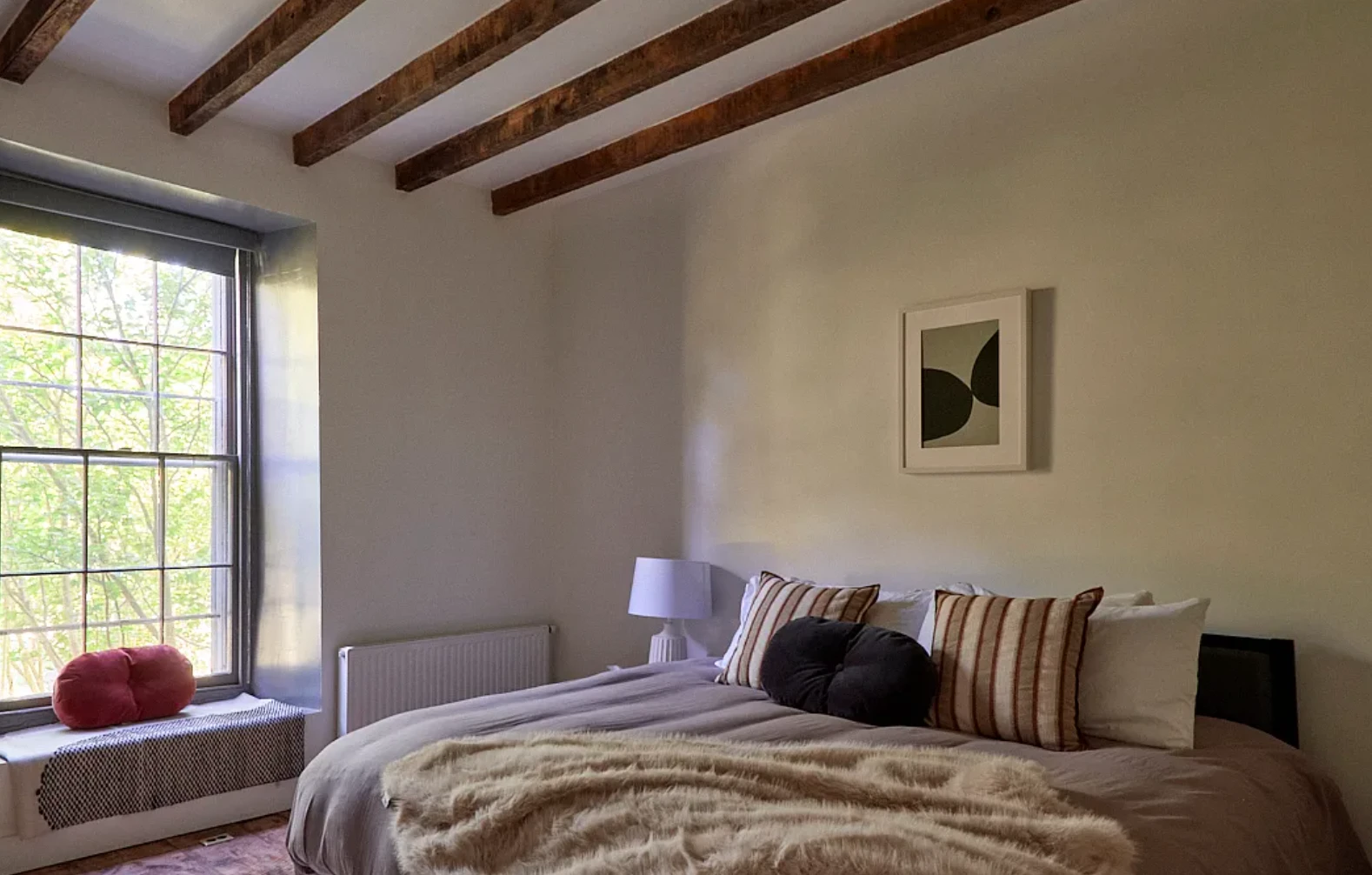 A sunlit bedroom with exposed wood beams, striped linen pillows, and a window looking into the trees